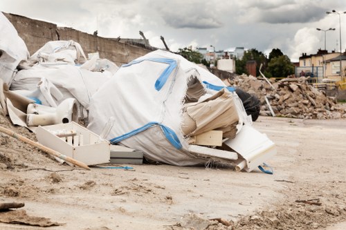 A large pile of mixed waste materials, including white plastic pipes, panels, and packaging, is gathered on a dirt surface in an outdoor area. The waste appears to be construction or renovation debris, with some items partially covered by white and blue protective sheeting. Surrounding the pile, the background features a rough, uneven ground with scattered dirt and small rocks, and a distant view of a stone retaining wall, a partially cleared site with loose soil and debris, and a line of trees and low-rise buildings under a cloudy sky. The scene indicates a site where rubbish removal services by Business Waste Removal Hendon could be required for construction waste clearance, situated in the Hendon area within London, suggesting the postcode range NW4 or NW11, consistent with the page titled 'Hendon Business Waste Removal'.