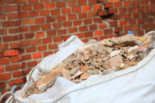 A large white bulk bag filled with mixed construction debris, including broken bricks, concrete fragments, and small pieces of tile or stone, placed outdoors on a paved area beside a red brick wall. The debris varies in colour from reddish-brown to grey and beige, with some pieces covered in dust. The bag is partially open at the top, with some loose materials spilling over the edges. The scene suggests an active rubbish removal process, typical of building or renovation waste clearance undertaken by Business Waste Removal Hendon, serving clients around the Hendon area in London. The environment appears well-lit under natural daylight, emphasizing the textures and colours of the debris and surrounding brickwork.