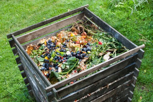 A large wooden skip situated outdoors on a grassy area, filled with a mixture of garden and household waste including various fallen leaves, grass clippings, fruit and vegetable scraps, small branches, and miscellaneous organic debris. The skip is constructed from dark, weathered wooden planks arranged horizontally, with a sturdy rectangular frame and open top. Surrounding the skip, there is lush green grass, and in the background, part of a garden or yard environment is visible. The image appears to depict a waste collection service, such as Business Waste Removal Hendon, efficiently managing organic garden and household rubbish outside a residential or commercial property in the Hendon area.