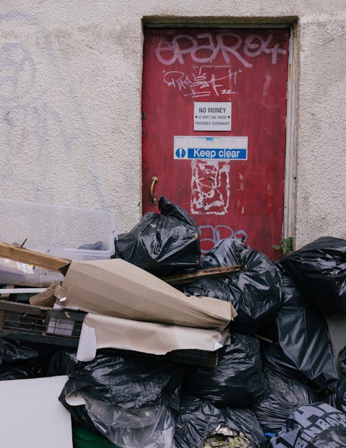 A collection of black waste disposal bags and discarded boxes and cardboard piled in front of a red metal door set into a textured, beige-painted wall. The door features graffiti tags and several stic